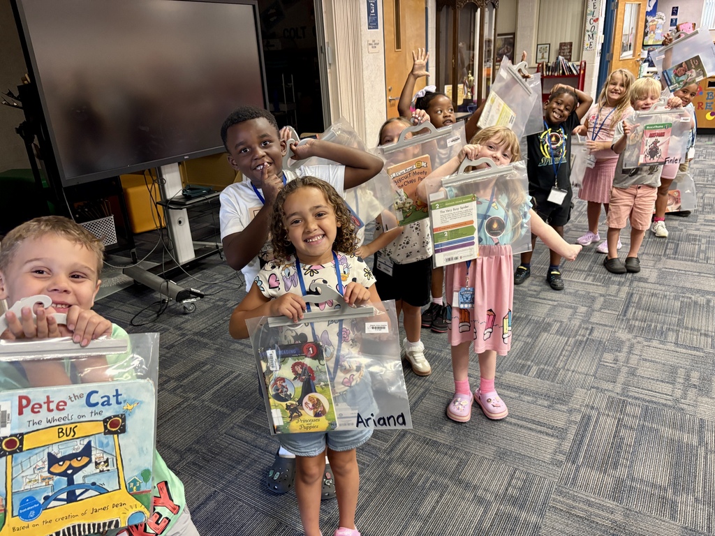 Several children in a classroom hold up clear bags containing books and toys. The room has carpeted floors, monitors, and doors.