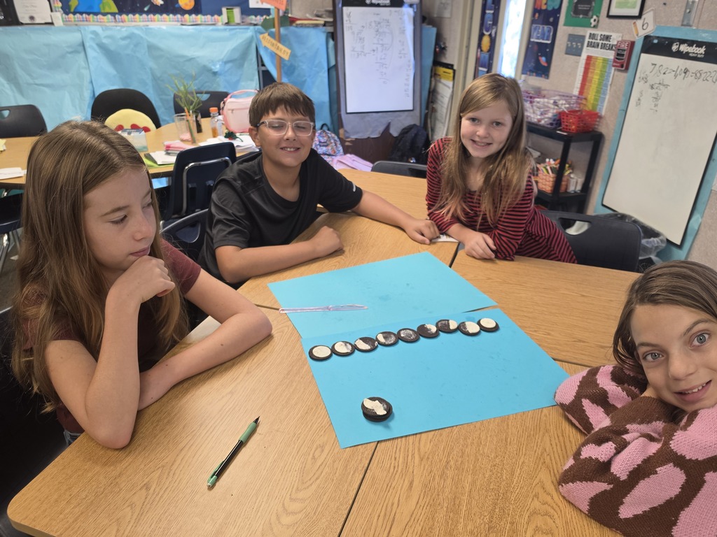 Four children are seated around a table, working on a project with a blue paper and small objects.
