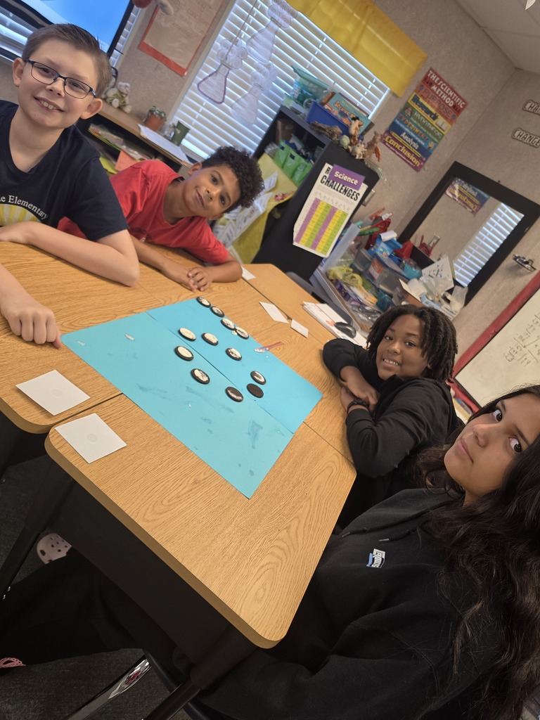 Four students are seated around a table, playing a game. The table has a blue board and counters.
