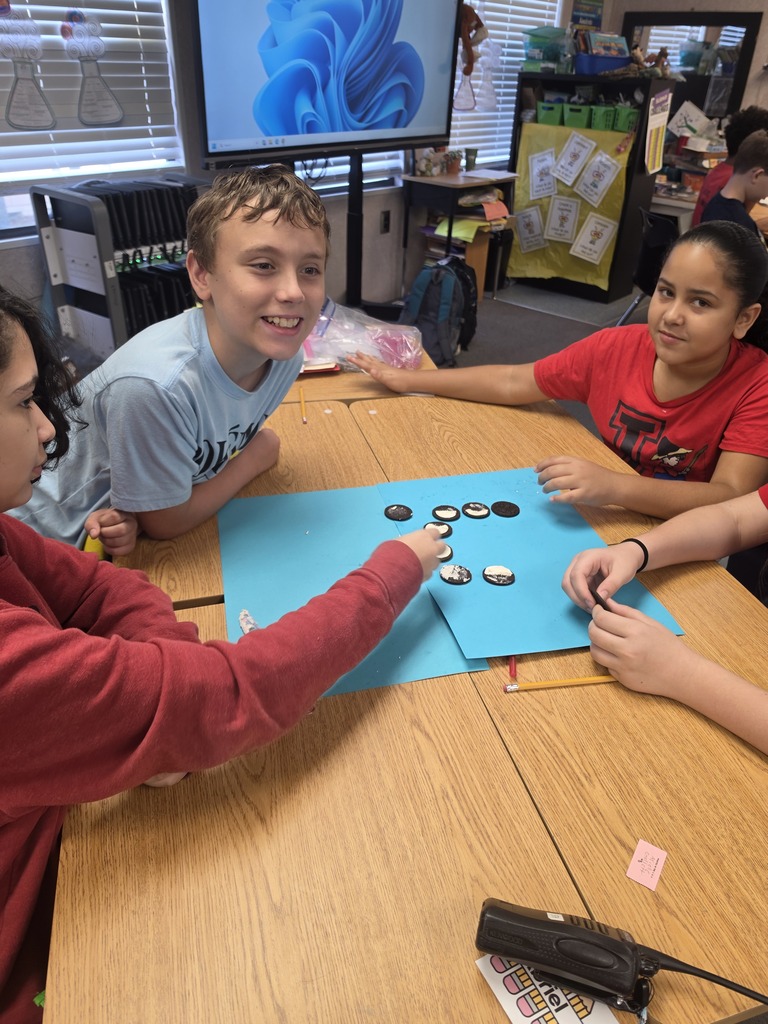 Four children are seated around a table, engaged in a game with blue paper and small objects.