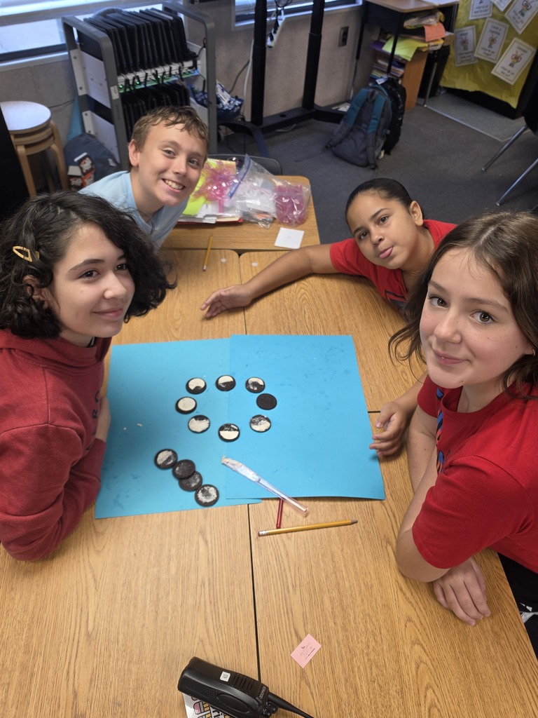 Four children sit at a table, smiling. Three of them lean over a blue paper with game pieces.