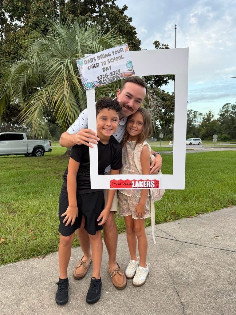 Family of three, with a boy, girl, and man, poses with a frame in an outdoor area.