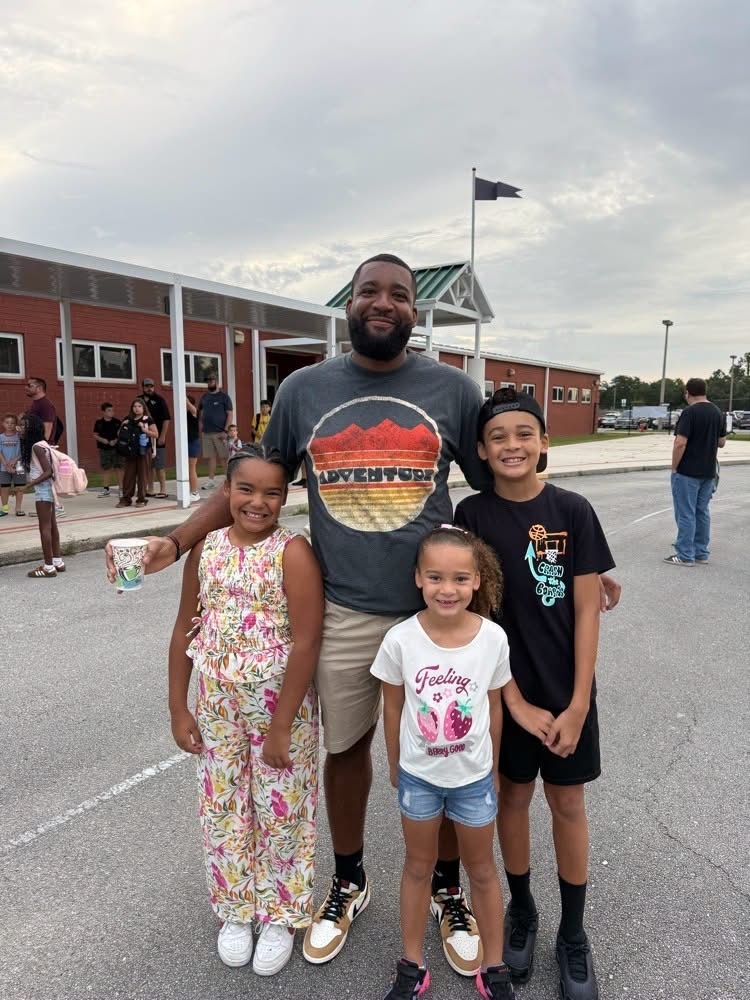 A man and three children stand together in a parking lot. The man wears a gray shirt with a design.