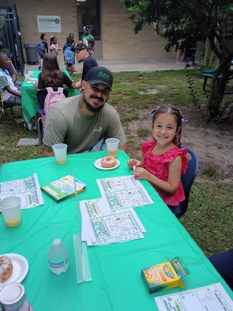 Man and girl at a table with puzzles and drinks. Trees and people in the background.