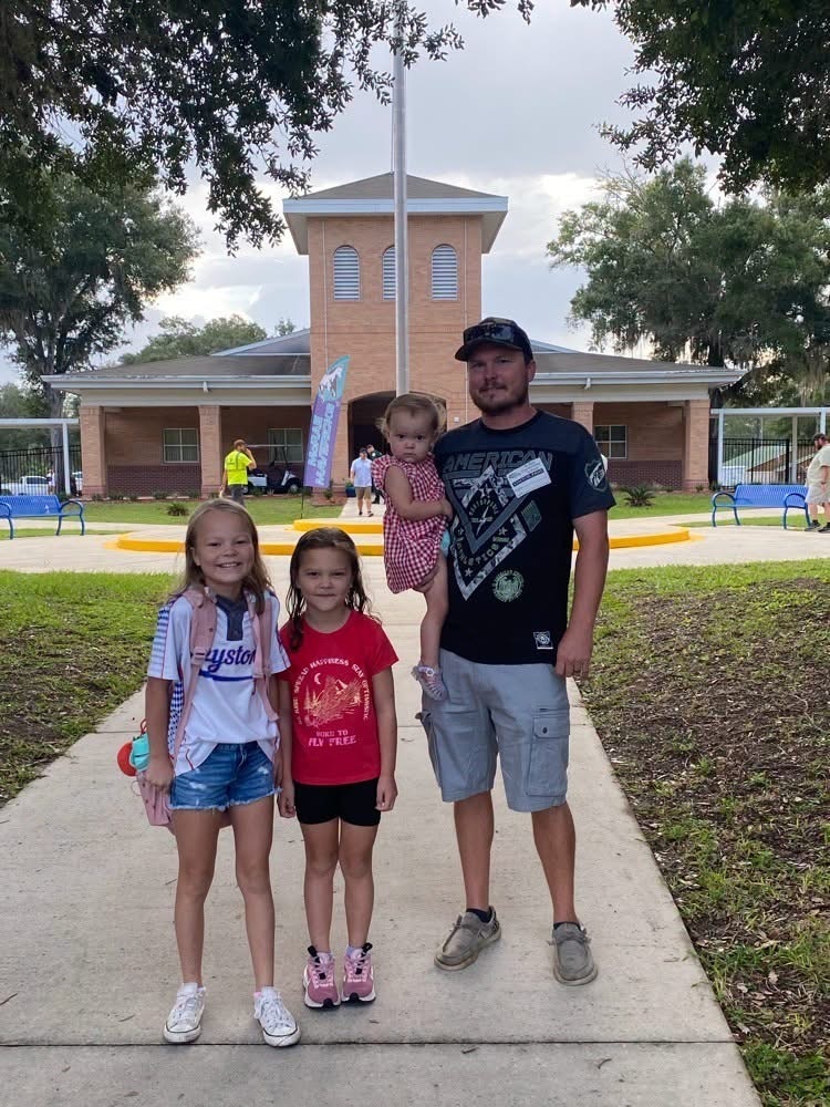 Family posing in front of a building with a flagpole. One child in pink carried by an adult.