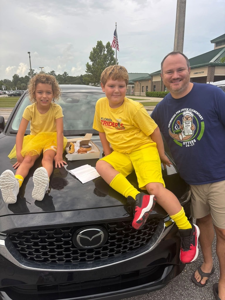 A man and two kids sit on a black car. The kids wear yellow. A box of food sits between them.