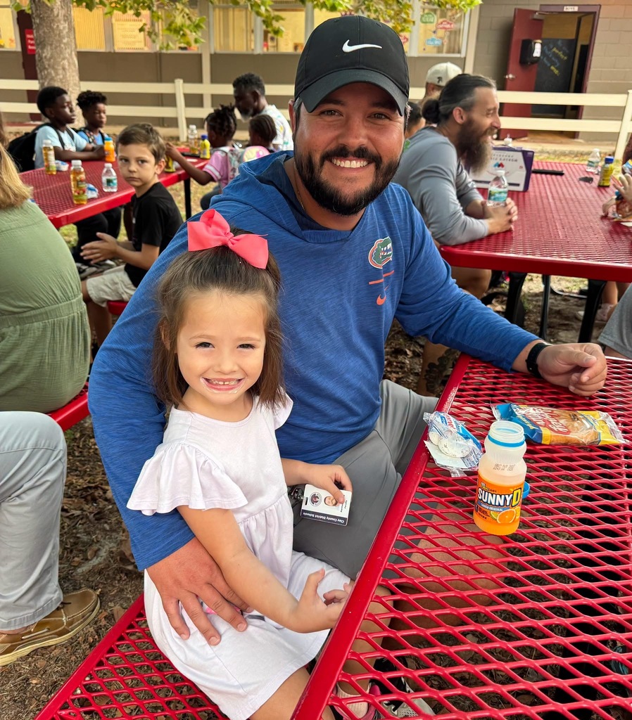 A man with a baseball cap and a girl with a pink bow sit at a picnic table.