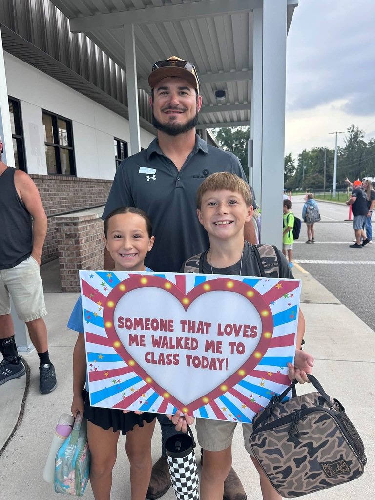 A man and two children pose with a sign in front of a building. The sign has a heart and says, "Someone That Loves Me Walked Me to Class Today!"