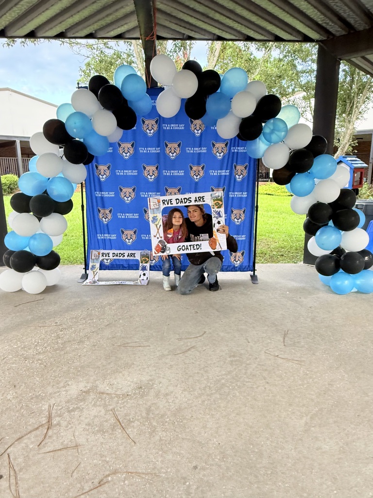 Two individuals crouch beneath a banner and balloons reading "Our Dads are GOATS". The banner is blue.