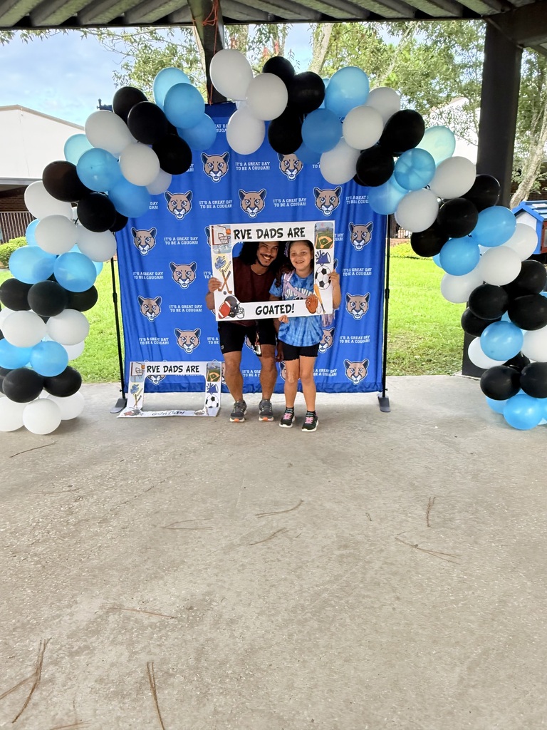 Two people stand under a balloon arch. One holds a frame with the text "Dads are". A blue backdrop with team logos is behind them.