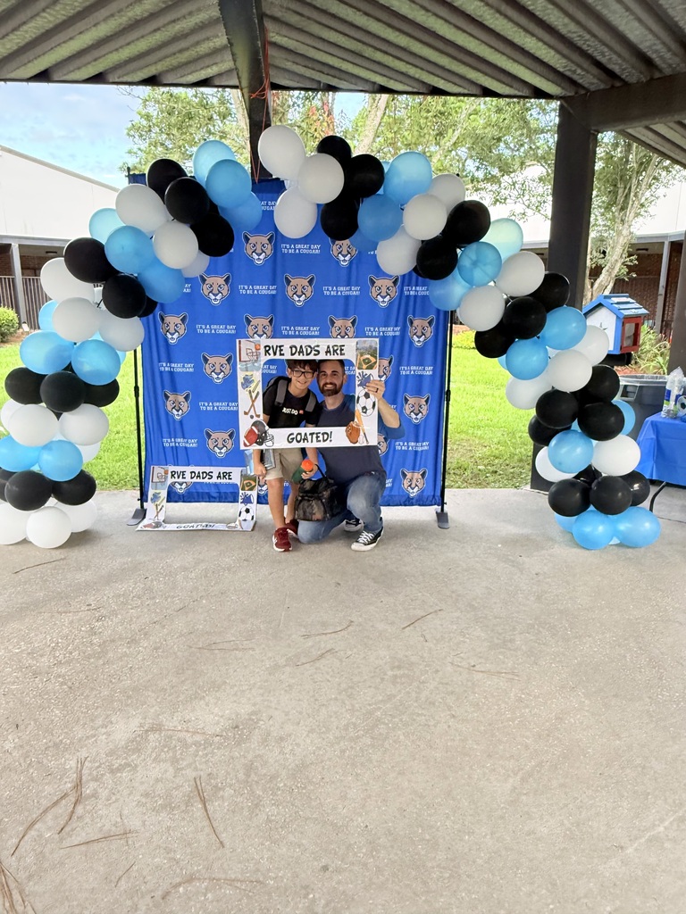 Two people pose under a balloon arch with a blue backdrop and white text. They sit on a concrete floor.