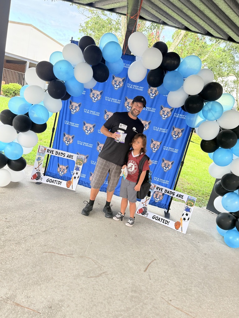 A man and a child standing under a balloon arch, with a blue backdrop displaying the word "Goats" and images of a cat.