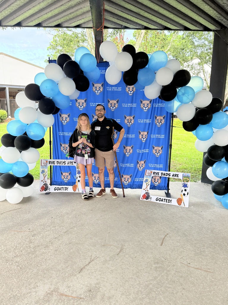 Two people, an adult and a child, stand under a balloon arch with a banner behind them.