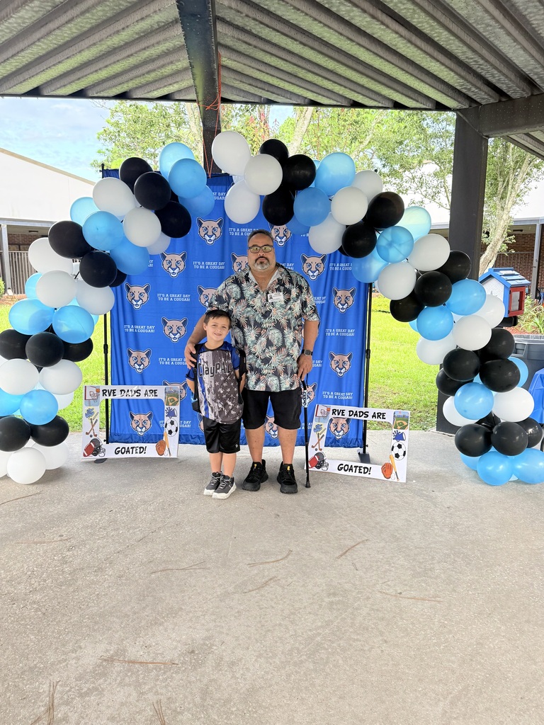 A man and a child stand under a large balloon arch with blue and black balloons.