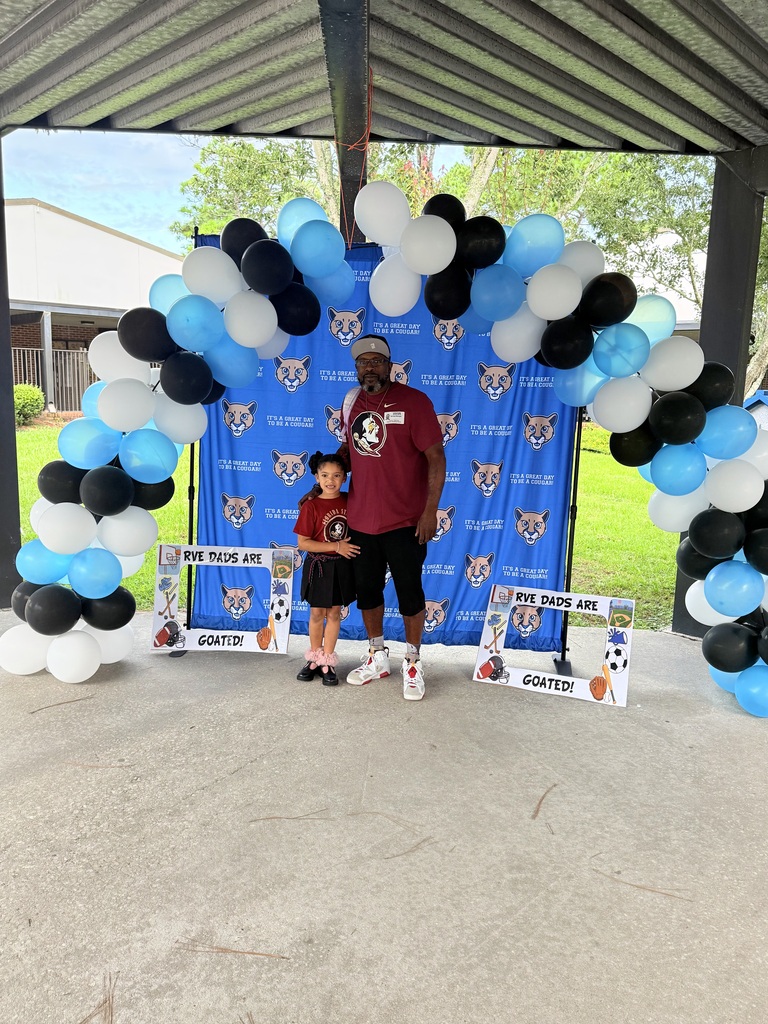 A man and a child stand before a blue backdrop with a balloon arch. They wear sports jerseys.