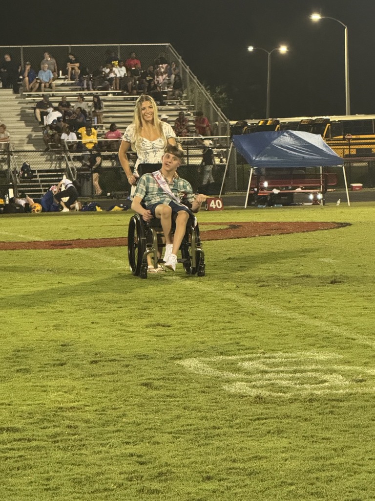 On a sports field, a woman stands behind a young boy in a wheelchair. Spectators are seated in the stands.