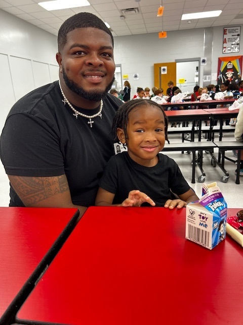 A man and a young girl sit at a table with a milk carton, smiling. Behind them, a school cafeteria is filled with people.