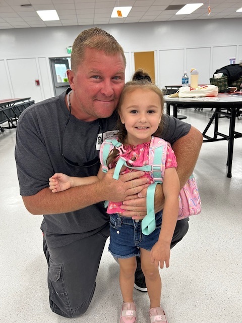An adult and child are smiling. The adult is kneeling and holding the child's backpack.