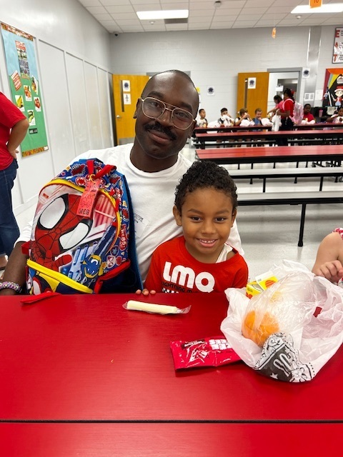 A man and a child sit at a table in a cafeteria. The child holds a backpack and snacks.