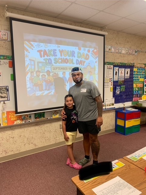 A man and a young girl stand together in a classroom. The man wears a hat and sneakers, the girl has pink shoes. Behind them is a projector screen.