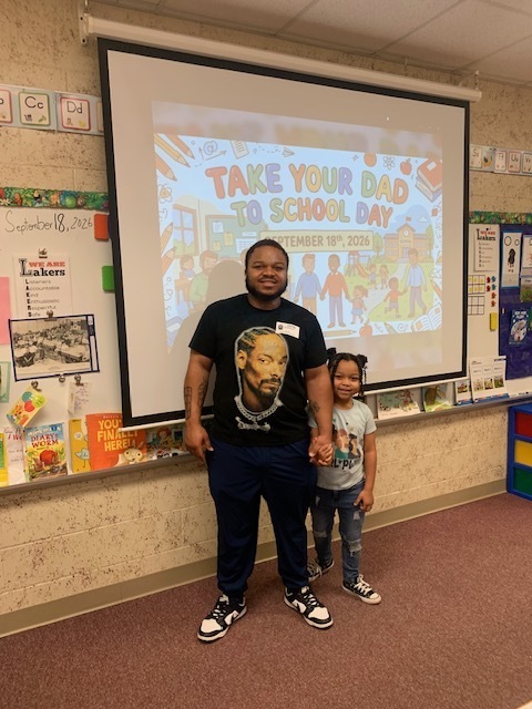 A man and a child stand in front of a projector screen with a message about Take Your Dad to School Day.