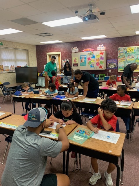 A classroom with children seated at desks. Some adults are assisting the students. The room has a projector and whiteboards.