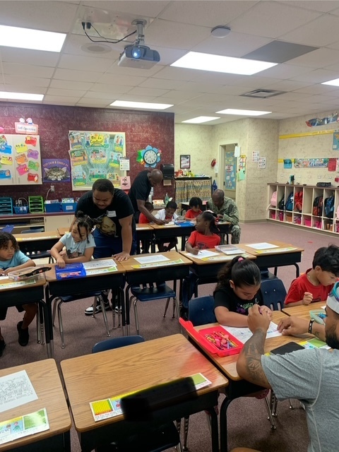 Classroom with students seated at desks. A teacher helps a student with a book. Bright lights and educational posters adorn the walls.