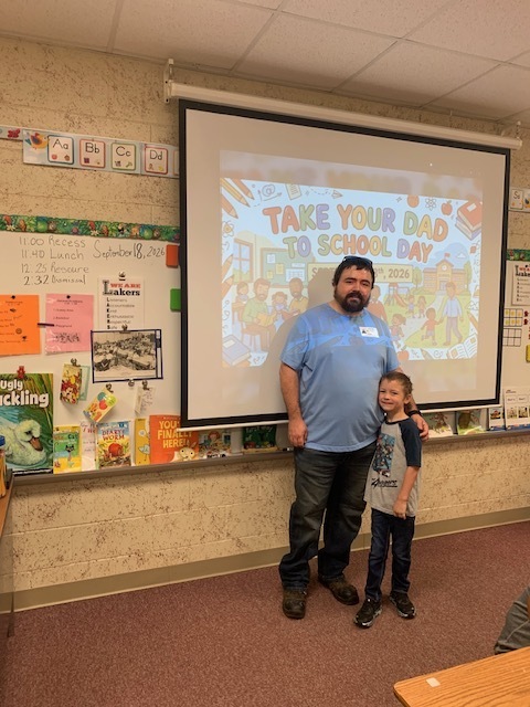 A man and a young boy stand before a projector screen displaying "Take Your Dad to School Day". The classroom background includes colorful posters and a bulletin board.