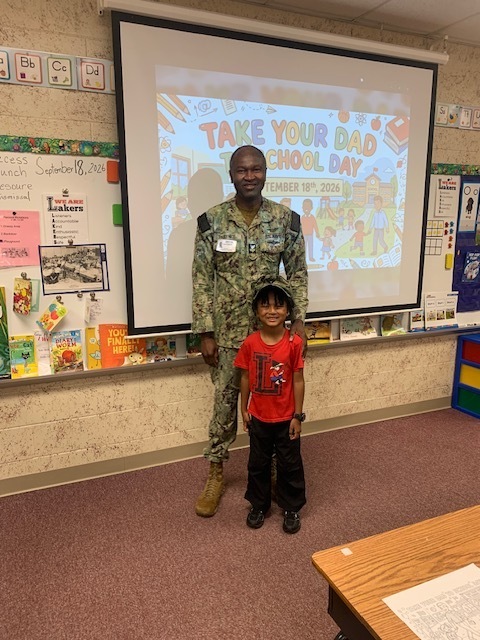 A man in military uniform stands next to a child. Behind them is a projector screen showing "Take Your Dad School Day."
