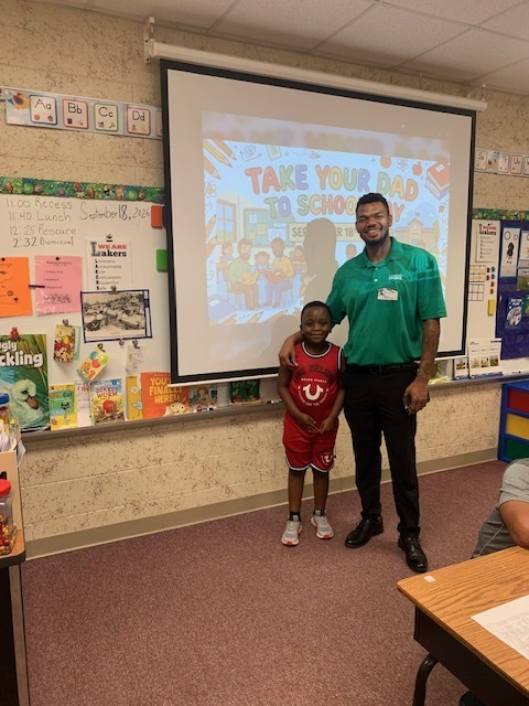 A man and a child stand in a classroom. The man wears green. They pose in front of a projector screen.