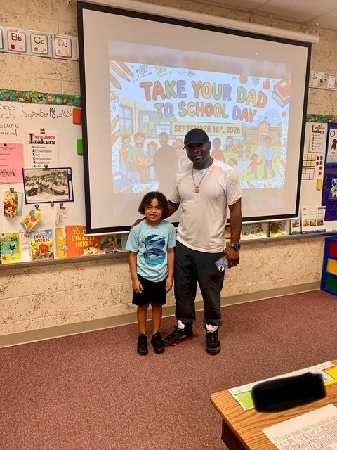 A man and a child stand in front of a projector screen that says "Take Your Dad to School Day".