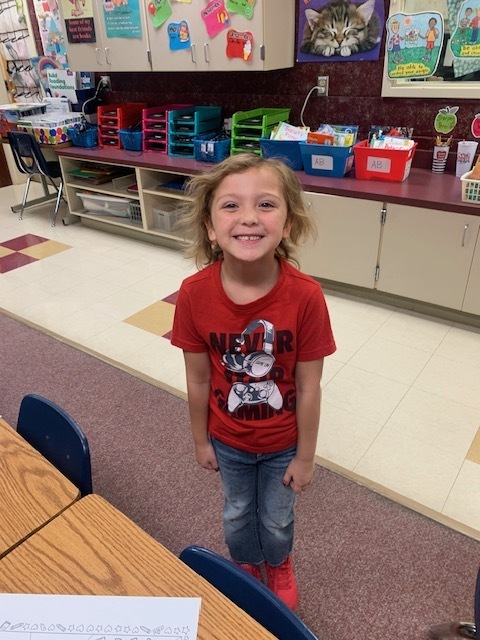 A child stands in a classroom with a bright smile, wearing a red shirt, blue jeans, and red shoes.