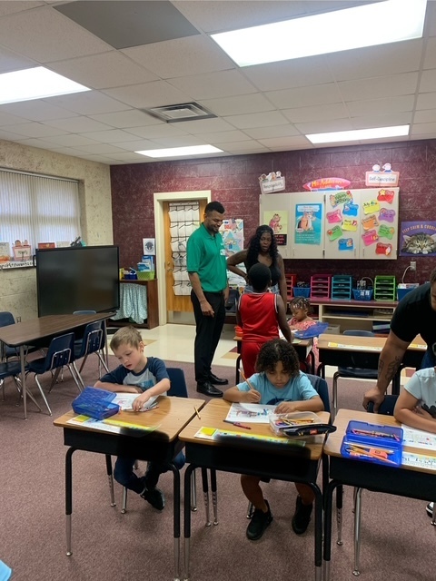 Kids sit at desks in a classroom. A teacher helps them. The room has red walls and white cabinets.