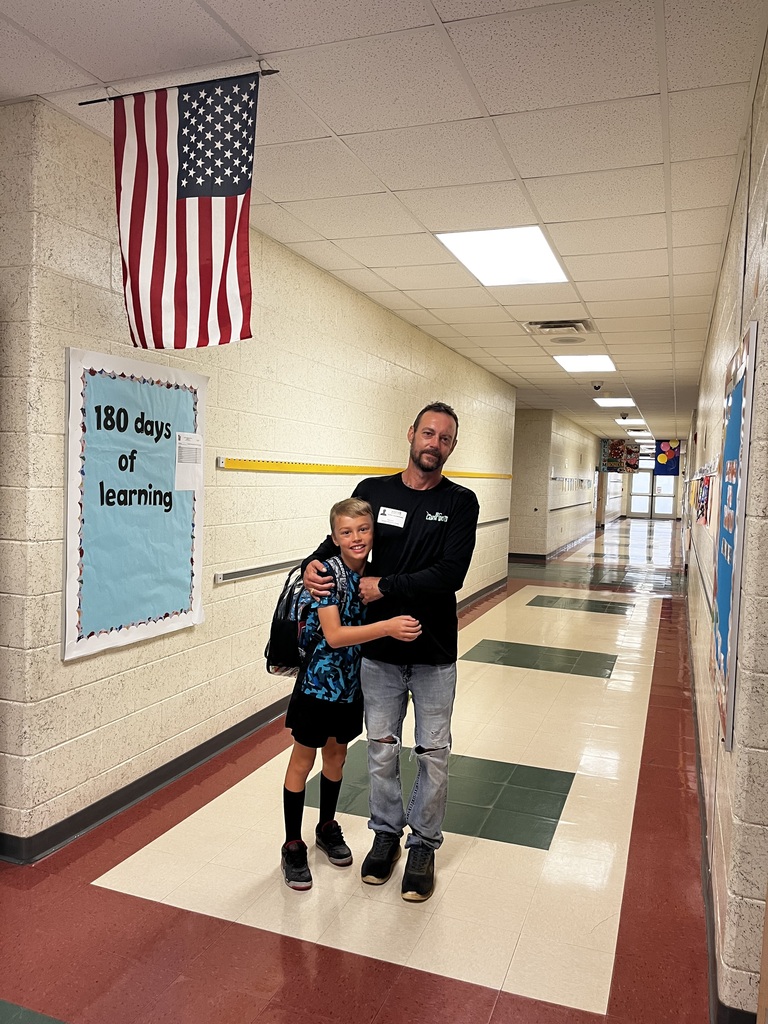 Man and child in a school hallway. They stand near an American flag and a sign that reads "180 days of learning."