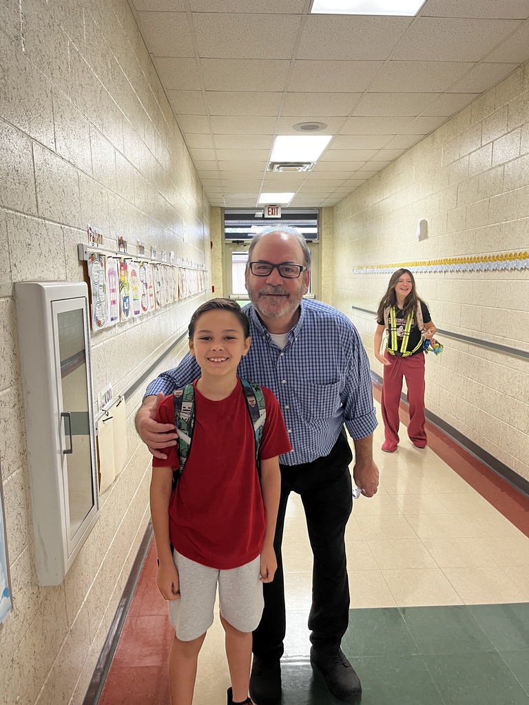 A man and a child pose in a school hallway. The child wears a red t-shirt and white shorts.