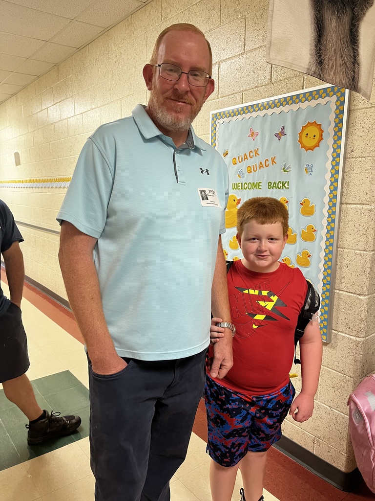 A man and a boy stand in a hallway. The man wears a blue shirt and jeans. The boy wears a red shirt and shorts. A banner hangs on the wall.