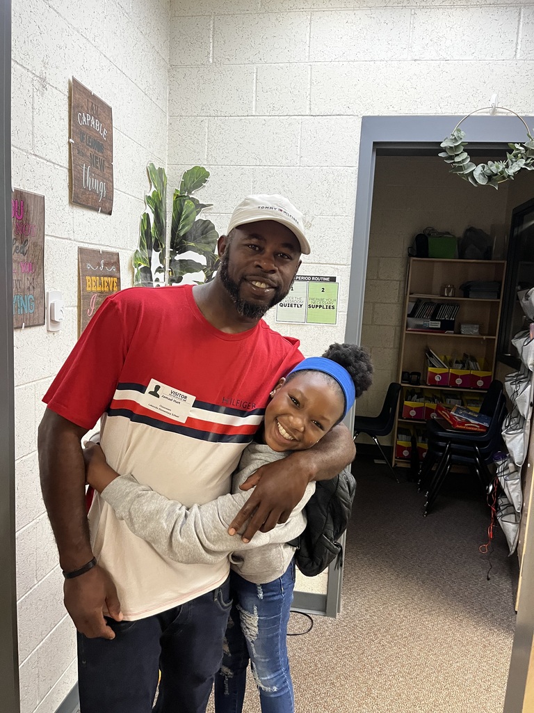 A man and a girl standing close, smiling, in a room with white walls, a door, and shelves with items.