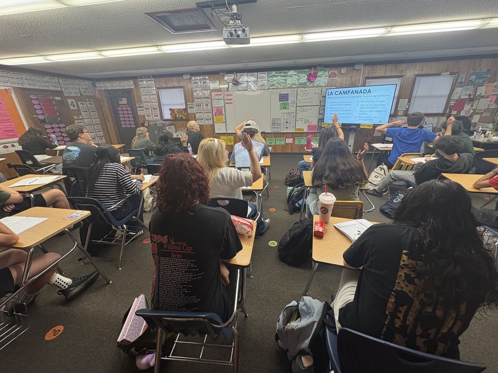 A classroom with desks and chairs, a projector screen, people sitting and some standing, one person raising their hand.