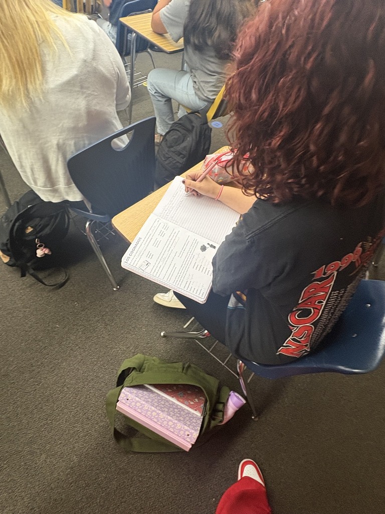 Students sit at desks in a classroom, one with red hair using a pen on paper, another with blonde hair.