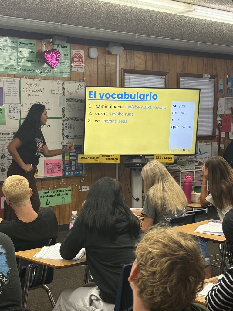 Classroom with woman at a projector screen reading El vocabulario. Students seated in chairs facing screen.