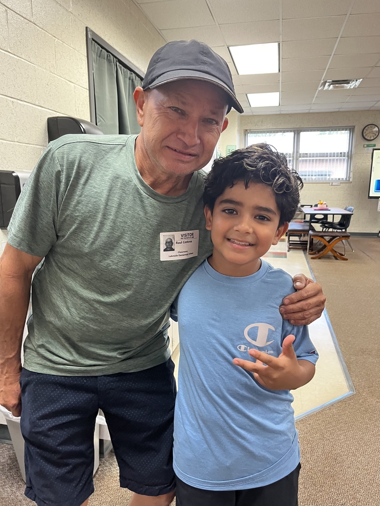 An older man in a cap and a boy in a blue shirt are standing and smiling in a room.