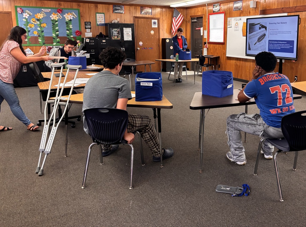 A classroom with two students seated; one has a cast on his leg. A teacher walks behind them.