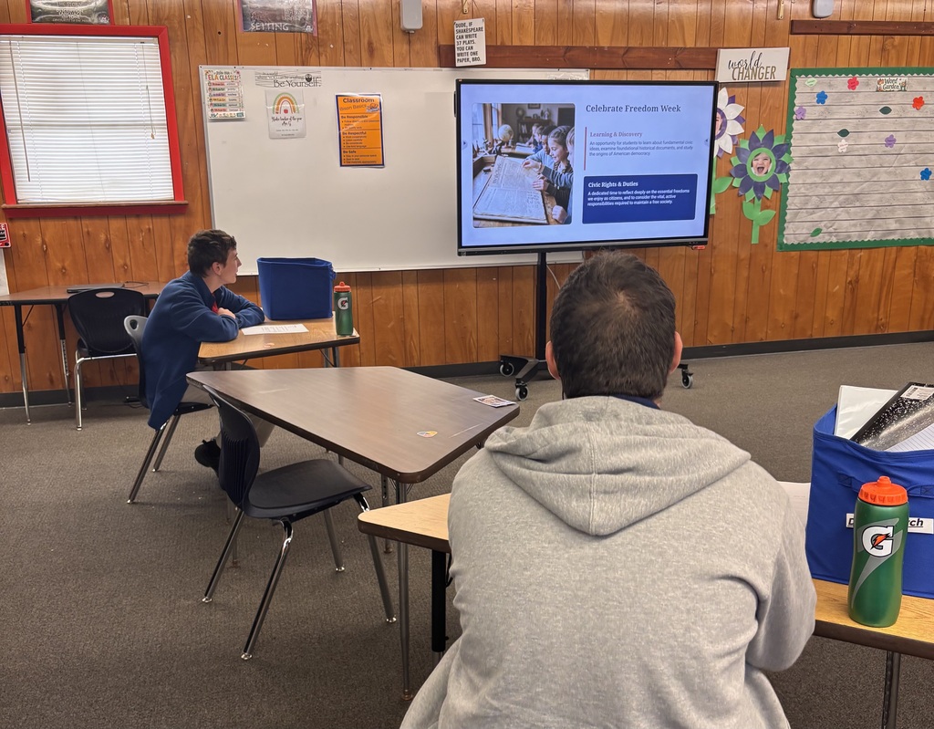 Two people sit at tables in a classroom with a monitor on the wall. One person wears a gray hoodie.