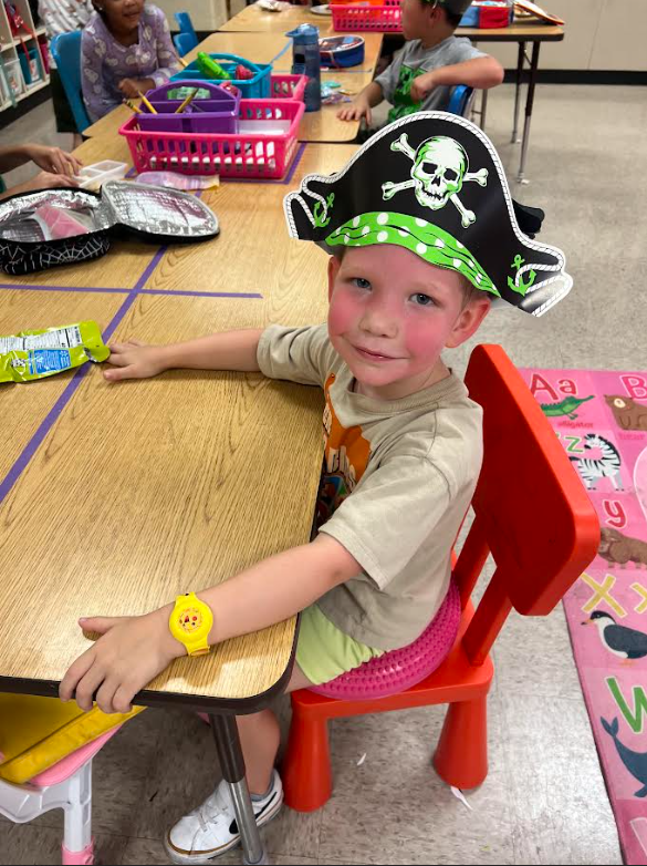 A young child wearing a pirate hat sits at a desk in a classroom. Desk has lunchbox and items.