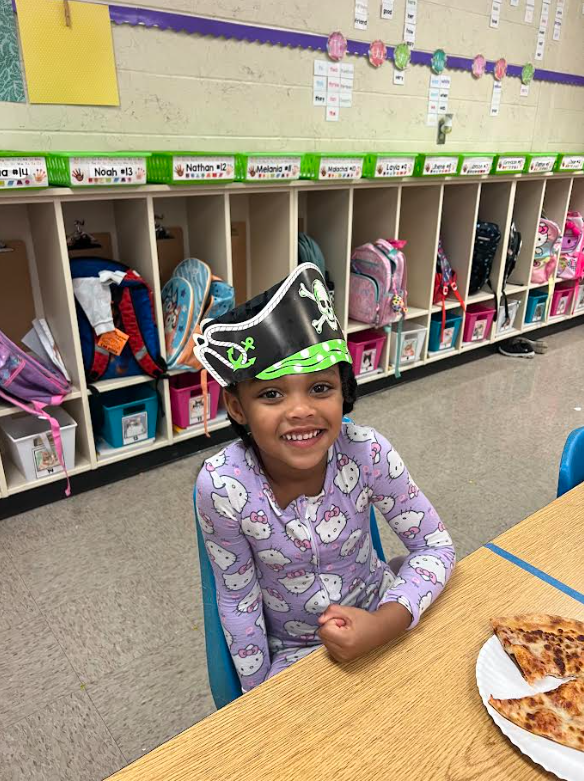 Child wearing a black and green pirate hat, smiling and seated at a table with a slice of pizza.