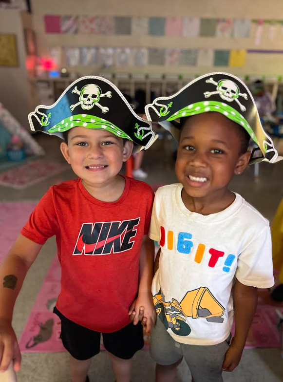 Two children in pirate hats smile at the camera. A red shirt and a white shirt are visible. Background includes pink carpets, a shelf, and other toys.