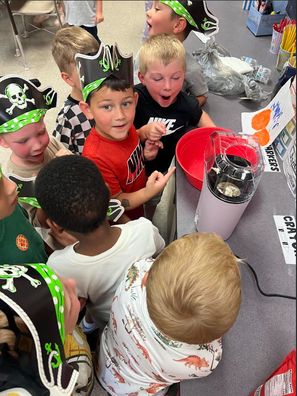 Children wearing pirate hats gather around a table, looking at a red bowl with a blender.