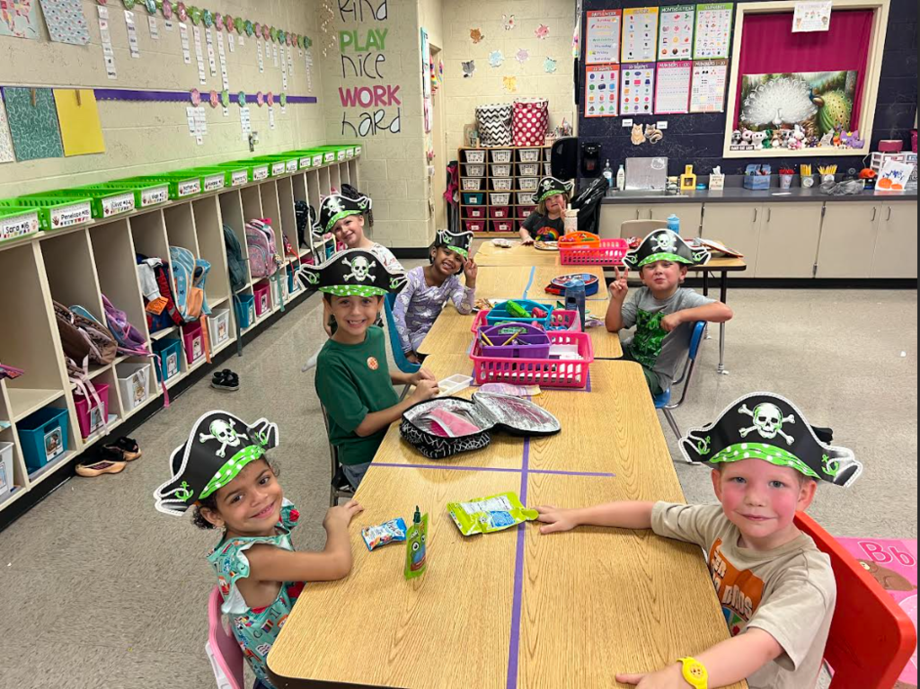 Kids in hats sit at a desk in a classroom with shelves, backpacks, and a bulletin board.