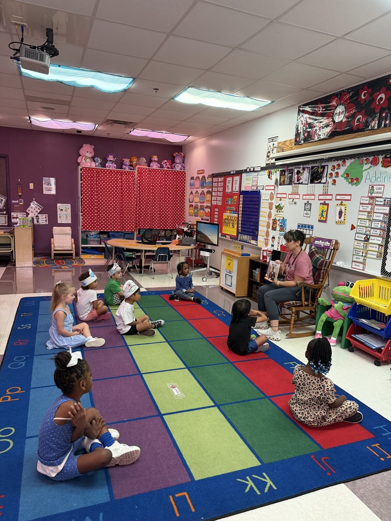 A classroom with a colorful rug. Kids sit on the rug and listen to a woman seated on a chair.