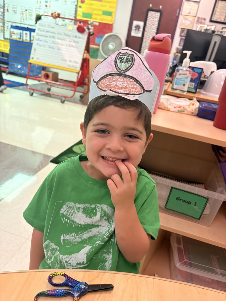 A young child in a green shirt and paper hat is smiling and sitting at a desk.